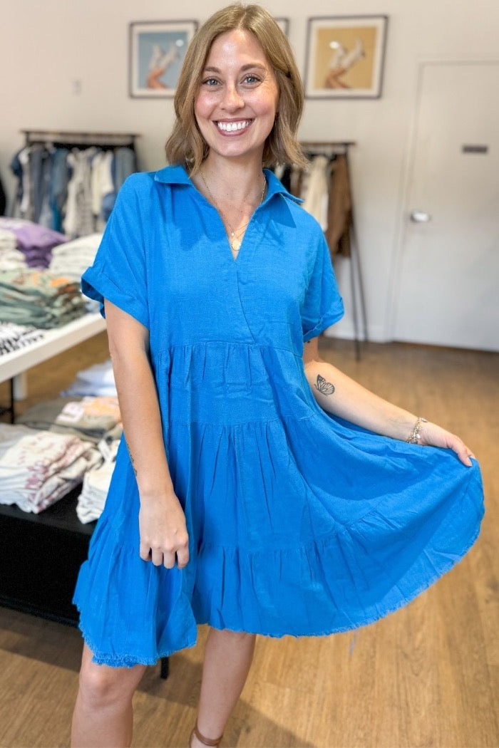 tiered flowy mini dress with a generous fit in a bright blue, collar with notch neck, rolled sleeve, and raw hem